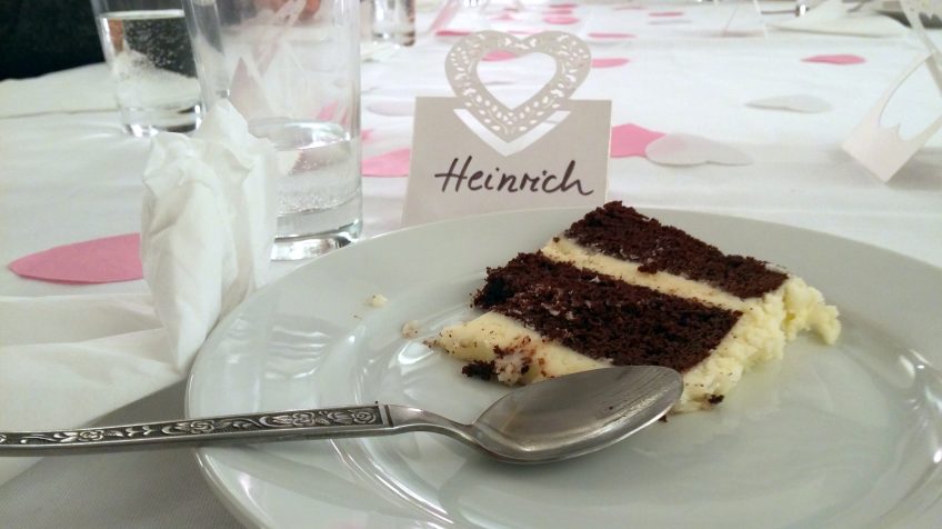 Ein Stück Hochzeittorte auf einem Teller. Links liegt ein Löffeln. Auf dem Tisch steht ein Namensschild, darauf steht "Heinrich".